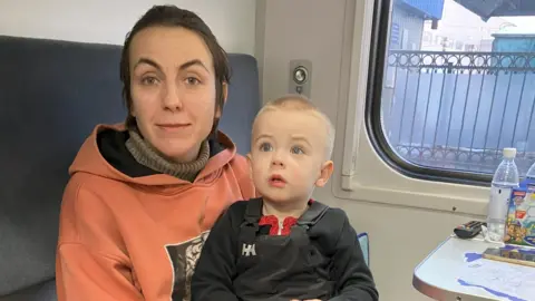 Alina sits on a seat in a train carriage with her son Taras on her lap. She is wearing a red top with hood while he is wearing a black top, possibly dungarees. There is food and drink on a small table to their right.