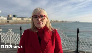 A woman wearing a red coat and red glasses. She is stood on a pier with the sea and seafront in the background.