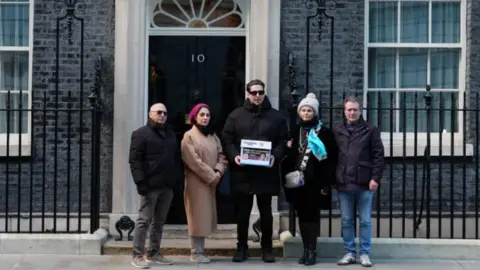 PA Media The group of five people stand wearing winter coats outside 10 Downing Street's door. The man in the middle who is wearing dark clothing is holding something that looks like a folder.
