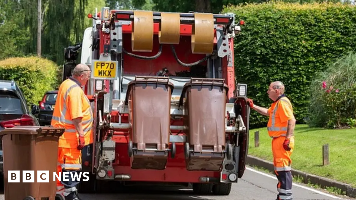 Two men in orange hi-viz overalls stand either side of the back of a waste collection vehicle. The truck is in the process of emptying two brown bins. Behind the van there is a patch of grass and several large green bushes, as well as a couple of parked cars on the road.