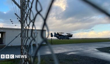 A military aircraft on a runway, photographed through a chain-link fence.