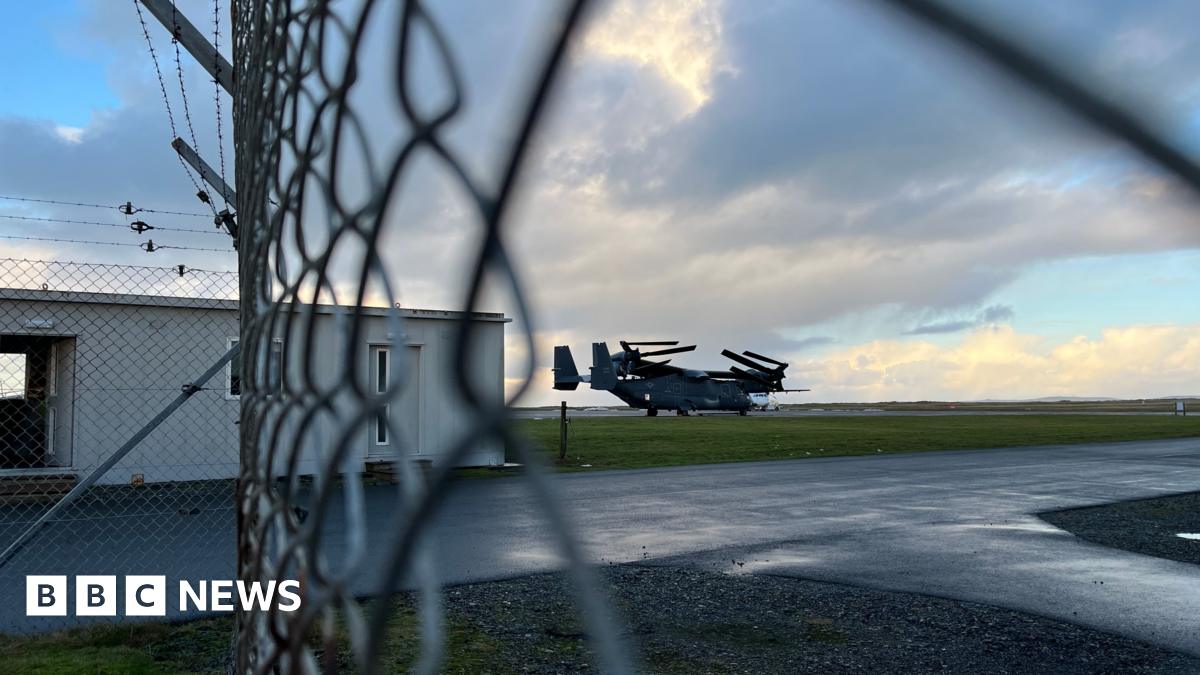 A military aircraft on a runway, photographed through a chain-link fence.