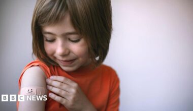 A young girl with brown, shoulder-length hair has rolled up the sleeve of her orange t-shirt to reveal a plaster with a smiley face on it on her upper arm, where an injection might go.
