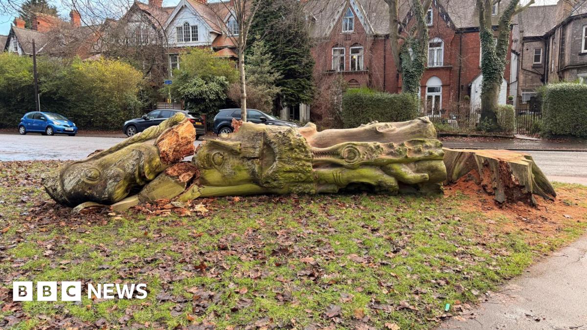 A decaying tree with a face carved into it is laid on its side after being chopped down. Residential houses can be seen in the background.