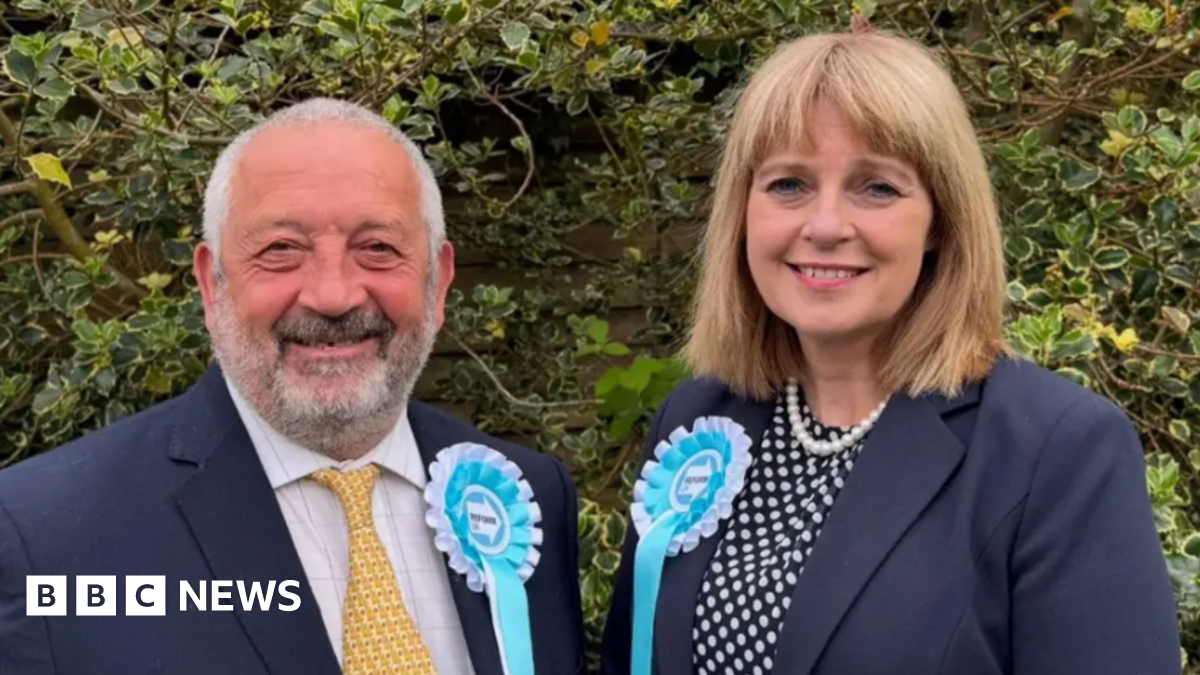 Marcus Lapsa and Jackie Gardiner look at the camera. They are wearing dark blue blazers and light blue and white Reform UK rosettes.