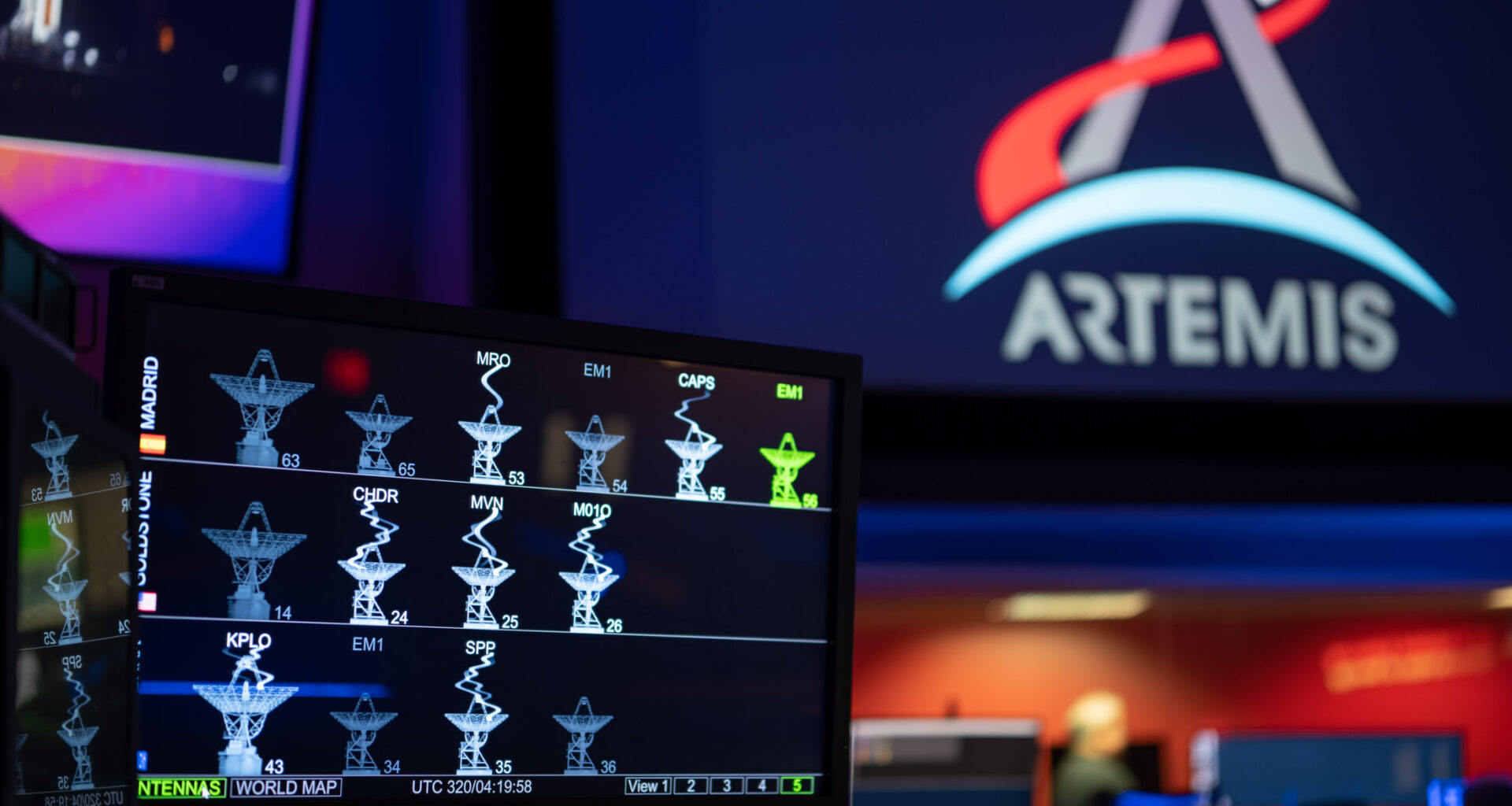 The DSN Now tool is displayed on a screen in the foreground—antenna images show real-time data provided by the Deep Space Network ground stations. The Artemis logo is seen in the background on a large screen.