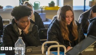 In a classroom, two students sit in their coats while working. One male student with black hair is leaning on his hand while in a puffer jacket. He is sat next to a female student who has long mousey brown hair who is writing with a pen on paper and is wearing a parker style coat.