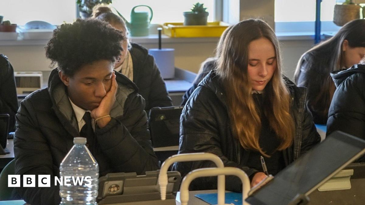 In a classroom, two students sit in their coats while working. One male student with black hair is leaning on his hand while in a puffer jacket. He is sat next to a female student who has long mousey brown hair who is writing with a pen on paper and is wearing a parker style coat.