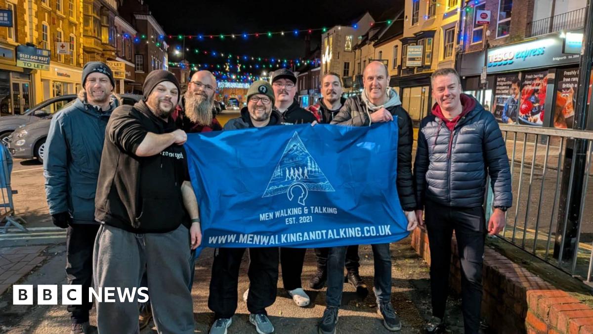 Men in a walking group stand in a shopping street and hold up a banner. It is night-time and there are Christmas lights above the road. Several men are standing near railings and they are wearing jackets, hats and trainers.