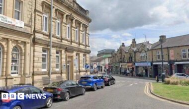 Four cars parked along a road in Brighouse, opposite a parade of shops.