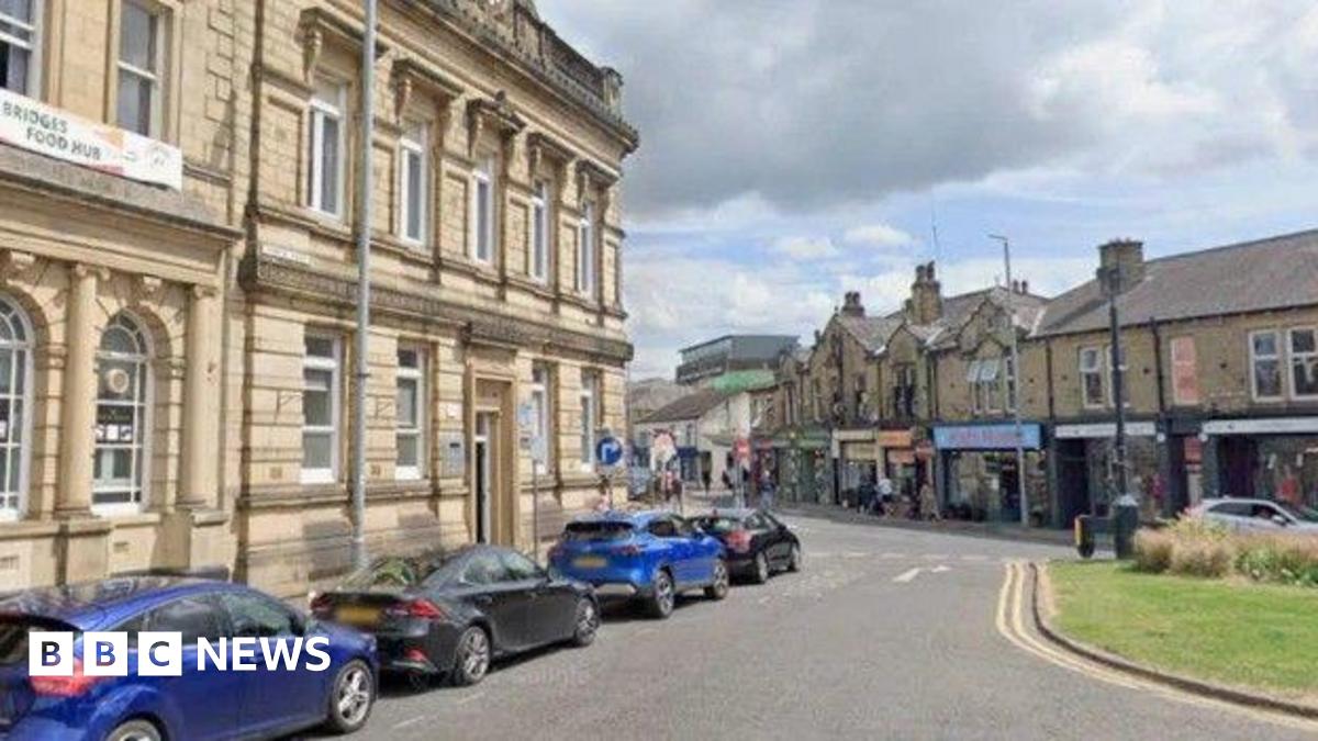 Four cars parked along a road in Brighouse, opposite a parade of shops.