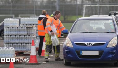 A man in his-vis clothing carrying packs of bottles of water in a car park. He is stood beside and looking into a blue car on the right, which is facing towards the camera. A woman is stood behind him, facing away, looking at a pallet stacked high with bottles of water.