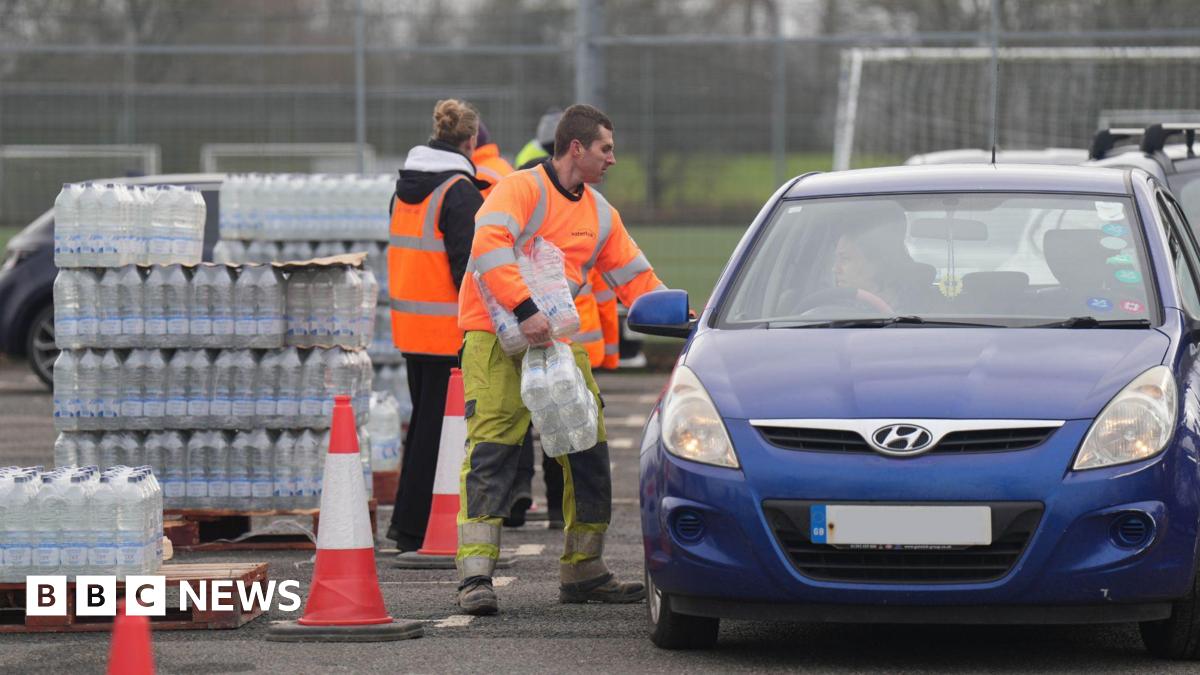A man in his-vis clothing carrying packs of bottles of water in a car park. He is stood beside and looking into a blue car on the right, which is facing towards the camera. A woman is stood behind him, facing away, looking at a pallet stacked high with bottles of water.