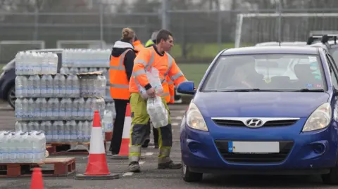 Eddie Mitchell A man in his-vis clothing carrying packs of bottles of water in a car park. He is stood beside and looking into a blue car on the right, which is facing towards the camera. A woman is stood behind him, facing away, looking at a pallet stacked high with bottles of water.