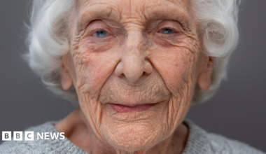 A head and shoulders portrait of Charlotte "Betty" Webb who has white hair and piercing blue eyes. she is wearing a light grey woollen jumper.