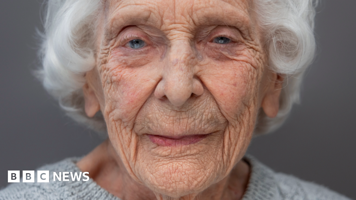 A head and shoulders portrait of Charlotte "Betty" Webb who has white hair and piercing blue eyes. she is wearing a light grey woollen jumper.