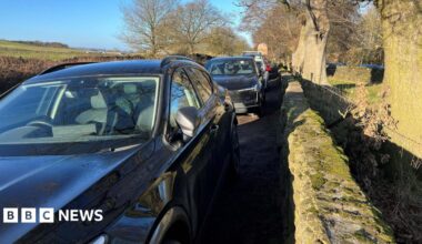 Millie Bobby Brown and David Harbour in character as Eleven and Jim Hopper. Eleven wears red shorts over grey leggings and a grey jumper. Jim Hopper wears a black leather jacket, a black beanie hat and dark trousers.They are speaking to director Frank Darabont on the set of Stranger Things. There is a vintage advertisement posted in the background with debrief on the ground.