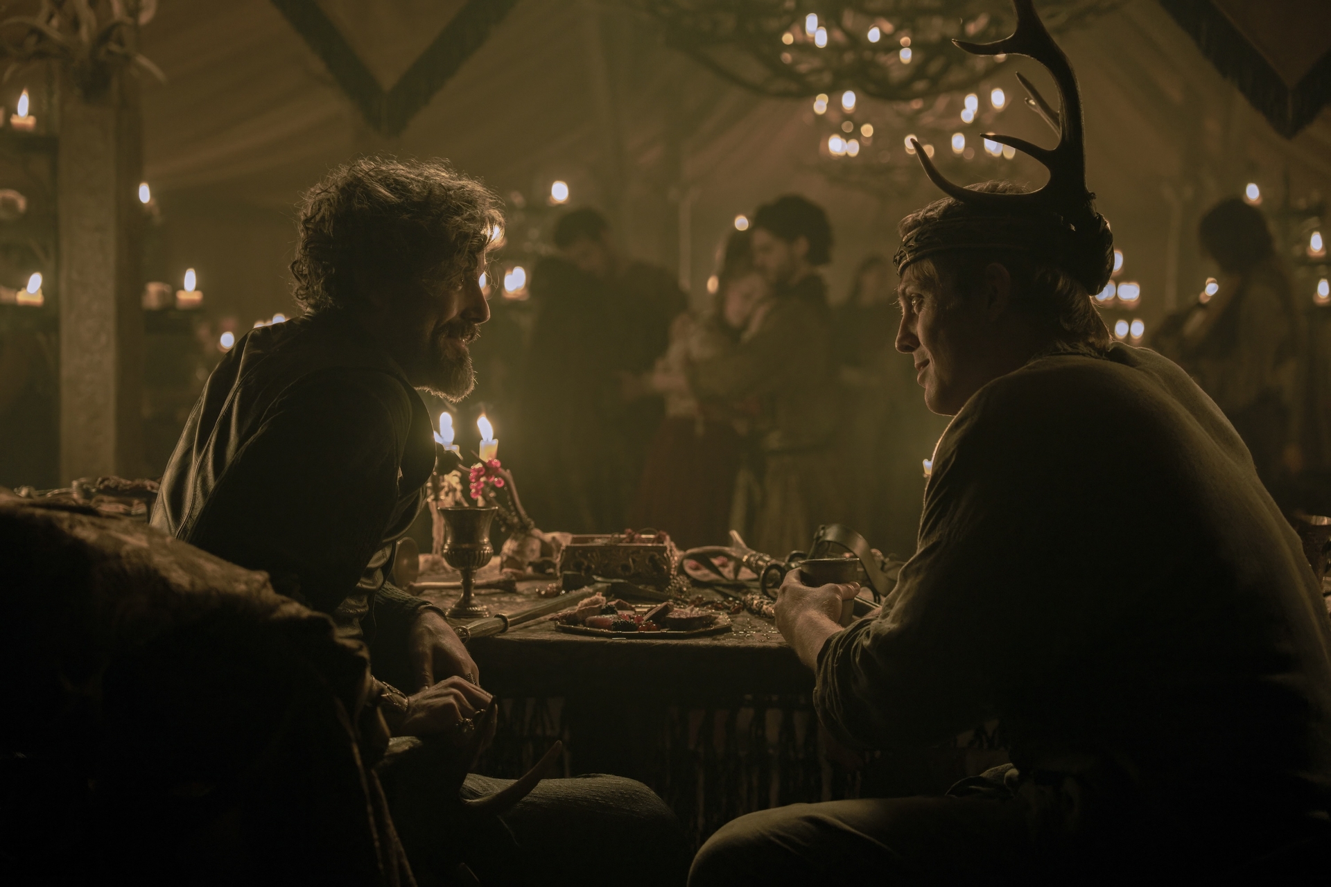 Two men sitting together at a table in room where a party is being held. The man on the right is wearing a crown adored with antlers.
