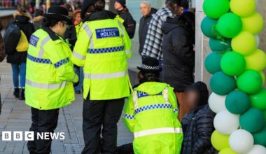 Police officers are talking to a man, whose face is blurred, sitting on a pavement outside some shops. Lime green, green and white balloons are next to him in a doorway.