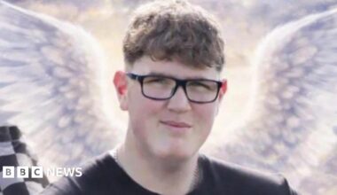 An image of a young man with glasses and short curly brown hair. He is looking at the camera and smiling. Behind him are some animated angel wings, a black and white chequered racing flag and a tyre.