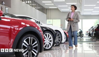 A woman wearing a grey jacket and blue jeans walks through a bright white car showroom, looking at a row of vehicles.
