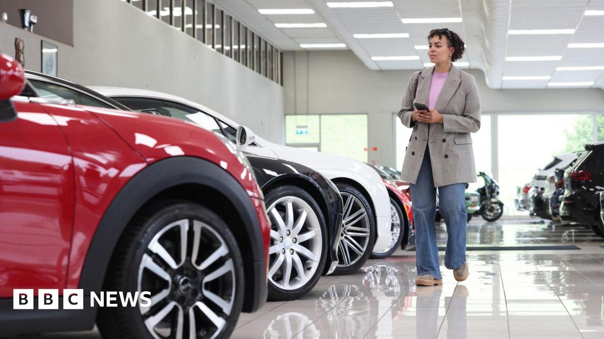 A woman wearing a grey jacket and blue jeans walks through a bright white car showroom, looking at a row of vehicles.
