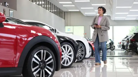 Getty Images A woman wearing a grey jacket and blue jeans walks through a bright white car showroom, looking at a row of vehicles.