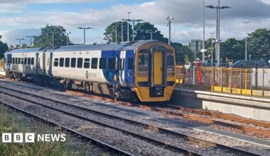 A blue and yellow Northern train at Ashington Station. It is a sunny day casting shadows across the three tracks. There is a workman in a hard hat and orange overalls leaning on railings on the platform.