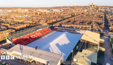 Aerial view of a football stadium with a protective cover, resembling a huge white tent, over the playing surface. There are four stands surrounding the pitch. The word "city" can be seen on one of the stands, which has red seats, and there are houses and a large cathedral in the background.