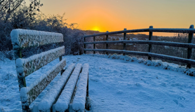 A park bench covered in snow  overlooking a snowed over fence while there is an organe glow in the sky and the sunsets.