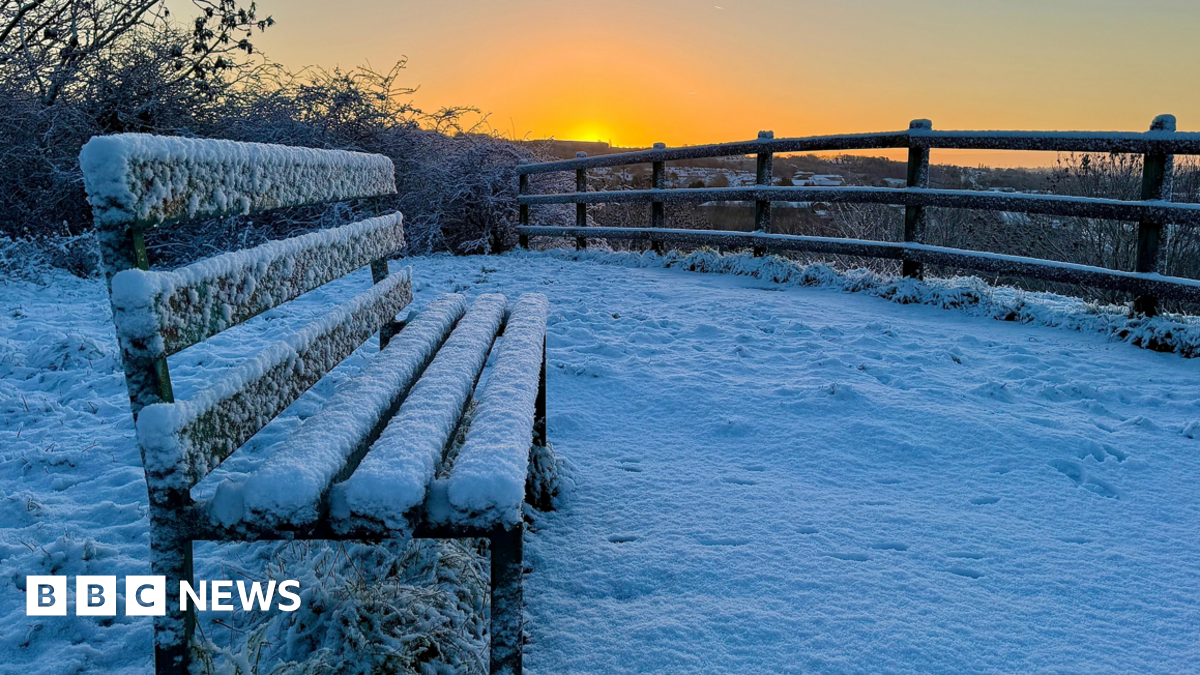 A park bench covered in snow  overlooking a snowed over fence while there is an organe glow in the sky and the sunsets.