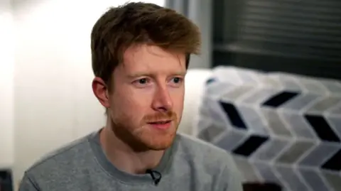 A man with short styled ginger hair and facial hair looks to his left. He is wearing a grey round neck jumper and has a clip mic on the collar