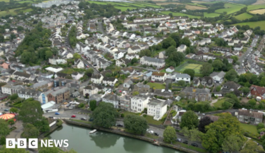 The picture shows an aerial view of a small town surrounded by rolling green hills and countryside. In the foreground, there is a calm river running horizontally across the bottom of the image, with a few small boats moored along its edge and a line of trees bordering the water. Behind the river, there are rows of houses and buildings packed closely together, forming a dense residential area. The houses are mostly two-storey, painted in light colours such as white, cream, and pastel shades, with sloping roofs.