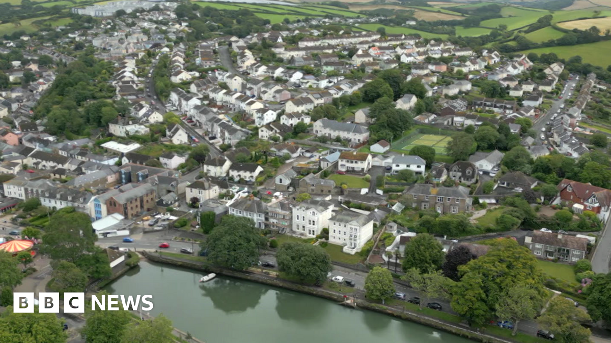 The picture shows an aerial view of a small town surrounded by rolling green hills and countryside. In the foreground, there is a calm river running horizontally across the bottom of the image, with a few small boats moored along its edge and a line of trees bordering the water. Behind the river, there are rows of houses and buildings packed closely together, forming a dense residential area. The houses are mostly two-storey, painted in light colours such as white, cream, and pastel shades, with sloping roofs.