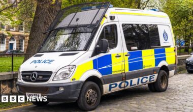 A police van with a riot shield attached to the windscreen. It is parked on a cobbled street.