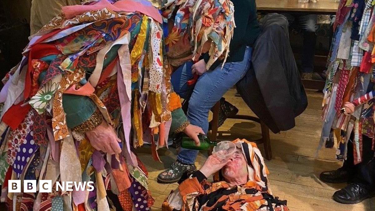 Three figures in coats covered in rags perform a play on a pub floor. One is laying on the ground with a funnel in his mouth while another pours liquid from a green bottle.