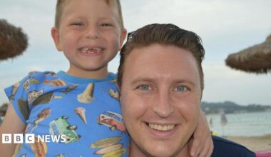 Image of a young boy with a blue t-shirt with images of camper vans and ice creams with his arm around a man with blue eyes and short brown hair.  The boy has gaps in his teeth but he and the man are smiling at the camera. In the background can be seen sand, sea and beach umbrellas.