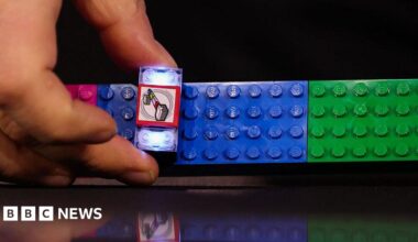 A close-up of a hand constructing a lit-up Lego Smart Brick on a set of pink, blue and green Lego blocks.