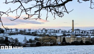 A lovely snowy scene with trees and a large building seen with a dusting of the white stuff. The sky is mostly clear with a few white and grey clouds moving away.