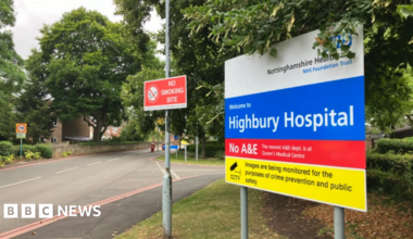 Millie Bobby Brown and David Harbour in character as Eleven and Jim Hopper. Eleven wears red shorts over grey leggings and a grey jumper. Jim Hopper wears a black leather jacket, a black beanie hat and dark trousers.They are speaking to director Frank Darabont on the set of Stranger Things. There is a vintage advertisement posted in the background with debrief on the ground.