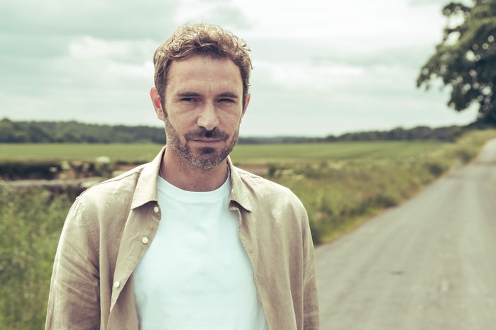 Oliver Farnworth as John Sugden for Emmerdale looking into camera in a white T-shirt and brown jacket