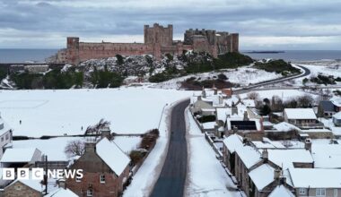 Bamburgh Castle rises above a snowy Bamburgh village. A winding narrow road leads towards the castle with the North Sea visible in the distance.