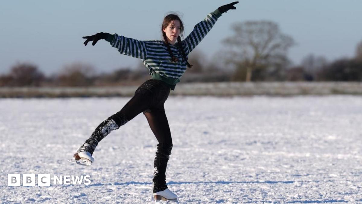 A woman is skating on an icy field. She has long, dark hair in plaits and is wearing a stripey top and trousers. She is a figure skater and her arms are elegantly outstretched as she skates with one foot on the ice and the other raised