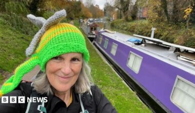 A woman with grey hair standing on a canal path wearing a knitted hat that resembles an air ambulance. A canal boat is next to her.