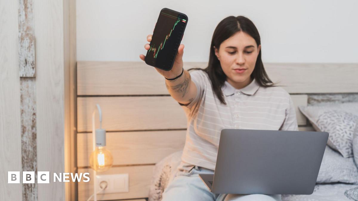 Young woman sitting on a bed with a laptop on her legs and holding out a mobile phone with a graph on the screen.