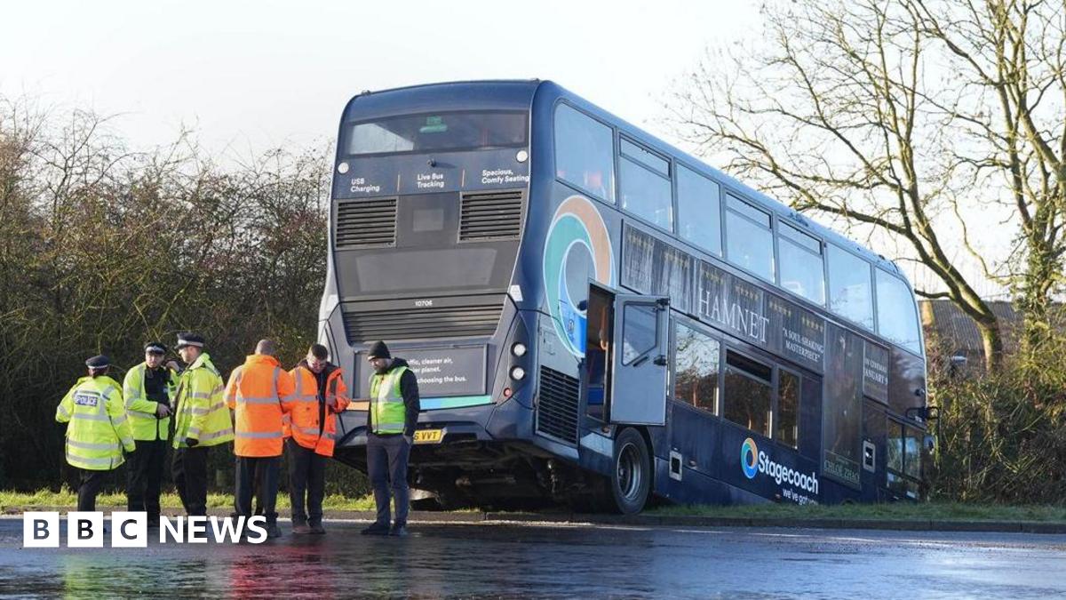 The Stagecoach bus is across a grass verge with its front wheels in a ditch and trees and houses in the background. Police are at the scene in hi-vis jackets and there are more people in orange jackets. The ground is wet.