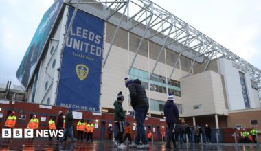 A landscape shot of Elland Road stadium's East Stand on a matchday. Banners with the club crest and name are visible. Three fans are seen at the front of the shot.