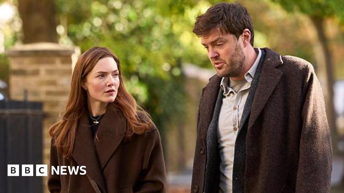 Holliday Grainger acting as Robin Ellacott and Tom Burke as Cormoran Strike in BBC drama Strike. Both actors are walking outside and are wearing brown coats. They are not looking at the camera.