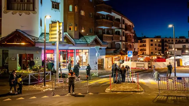 Police and press wait outside a cordoned off are in Crans-Montana. There's residential buildings and shops illuminated by night lights, white sheets and a red plastic tent in the background outside Le Constellation bar
