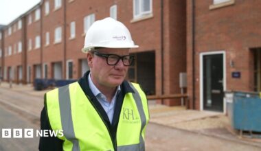 Richard Parker wearing a white builder's hat, glasses and a hi-vis jacket over a suit, stands and looks to our right. Behind him is a row of homes that are under construction.